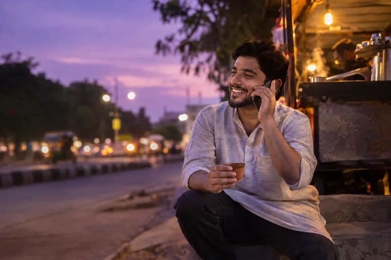 A young man laughing on a phone call at a chai stall in the evening